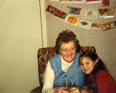 a lady sat in a chair, a child next to her, both smiling to the camera. both with glasses in hands. Christmas cards on hanging not the wall behind.